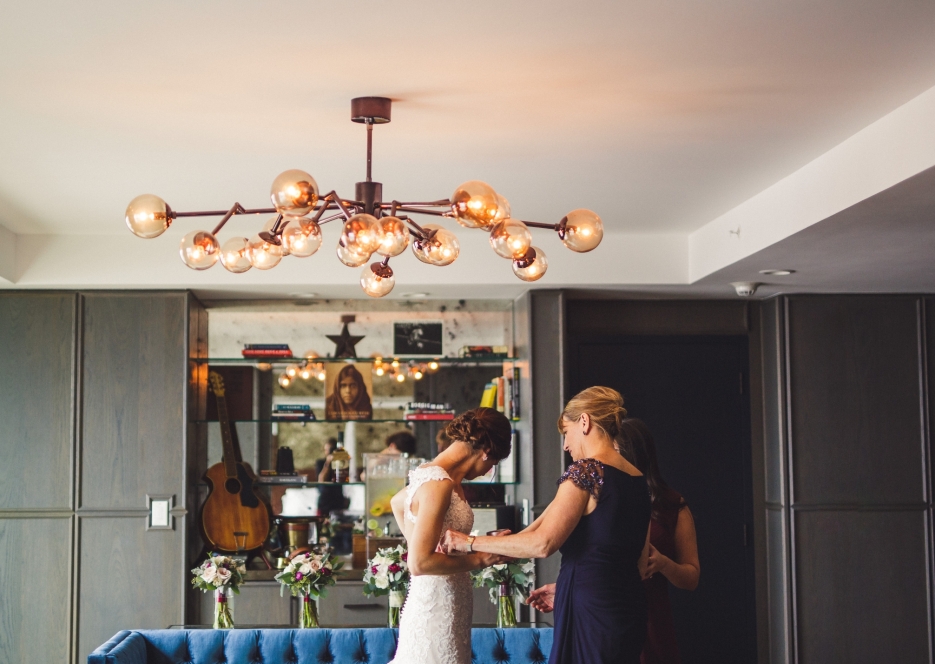 A woman in a white dress and another woman in a dark dress stand in a room with a blue couch, flowers, and a chandelier.