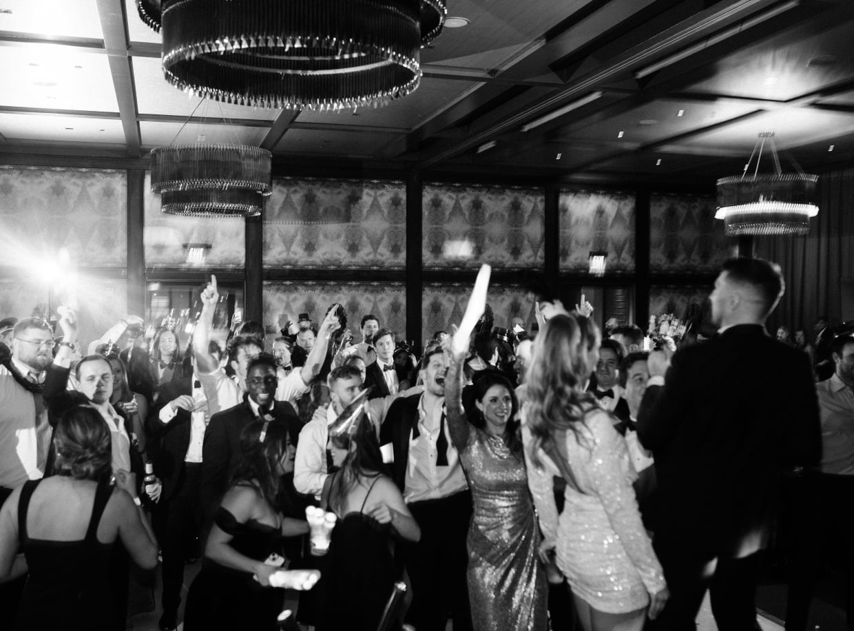 A large group of people in formal attire dancing and celebrating in a luxurious room with chandeliers.