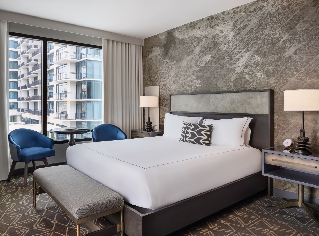 A hotel room with a large bed, two blue chairs, a window with a city view, and a bench at the foot of the bed.
