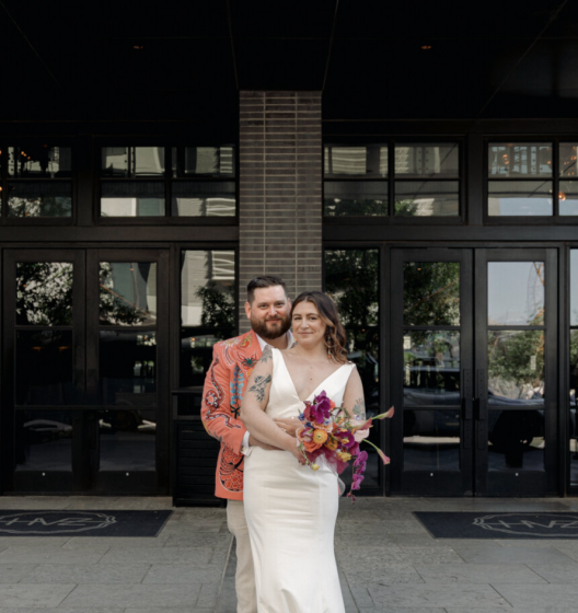 A wedding couple stands in front of a modern building with large glass doors and a brick column.