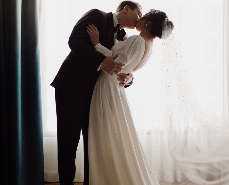 A romantic moment of a couple in wedding attire in front of a window.
