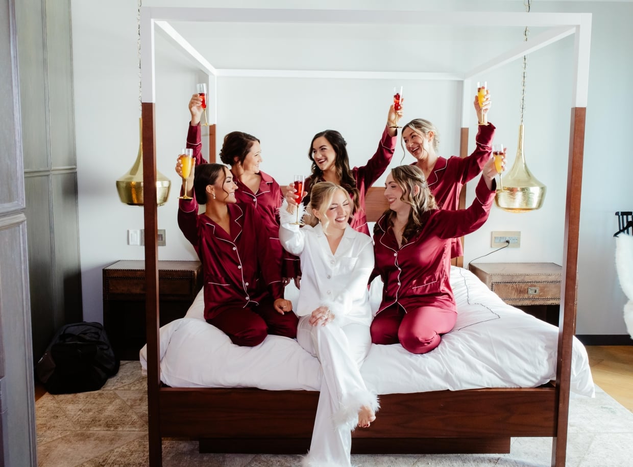 A group of women in red satin pajamas sit on a four-poster bed, holding glasses of champagne.