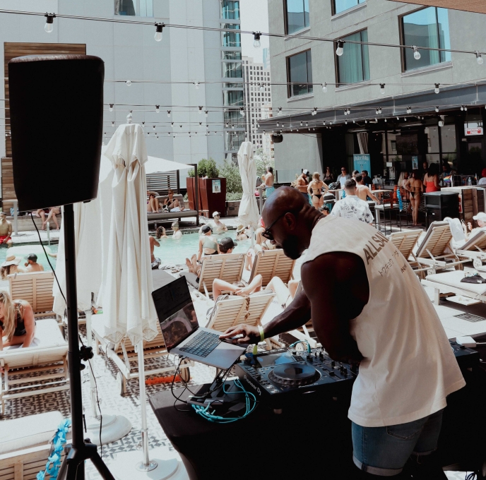 A man DJing at a poolside event with people lounging on chairs and swimming in the background.