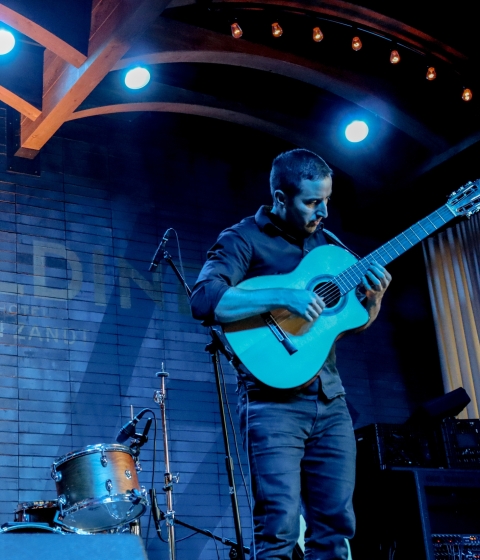 A man plays an acoustic guitar on a stage with a drum set and spotlights.