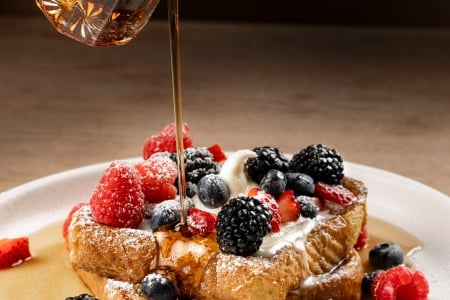 Maple syrup being poured over a golden Belgian waffle topped with a mound of fresh mixed berries and powdered sugar.