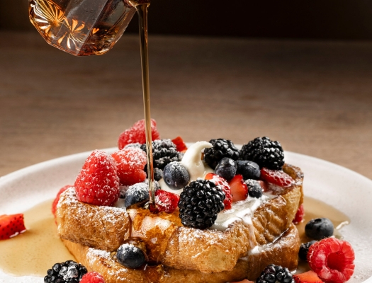 Maple syrup being poured over a golden Belgian waffle topped with a mound of fresh mixed berries and powdered sugar.