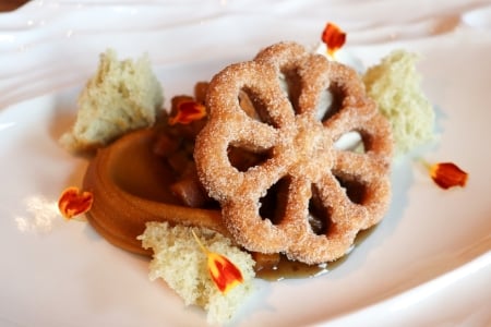 A close-up of a delicate, flower-shaped fried pastry, possibly a buñuelo, dusted with sugar and served with caramel sauce.