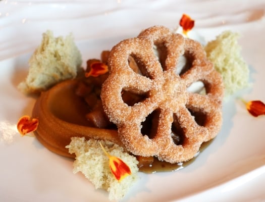 A close-up of a delicate, flower-shaped fried pastry, possibly a buñuelo, dusted with sugar and served with caramel sauce.