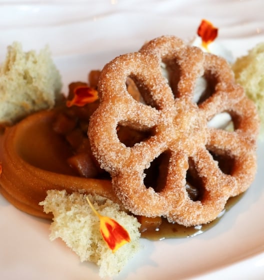 A close-up of a delicate, flower-shaped fried pastry, possibly a buñuelo, dusted with sugar and served with caramel sauce.