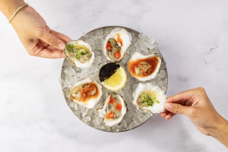 Two hands serve a platter of fresh oysters on the half shell, each with a unique, colorful garnish.