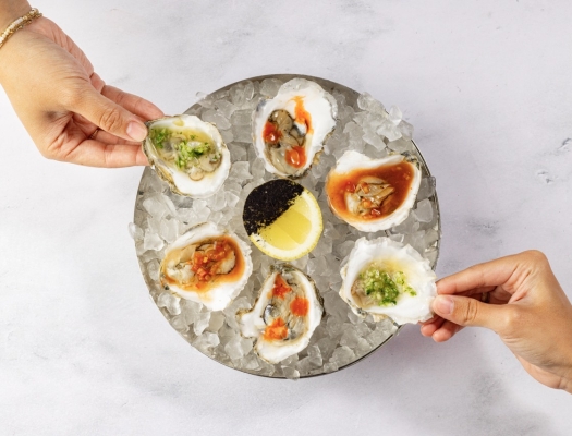 Two hands serve a platter of fresh oysters on the half shell, each with a unique, colorful garnish.