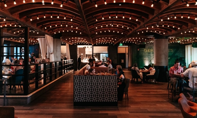 A large dining room with a high ceiling, wooden floors, and string lights, filled with people sitting at tables.