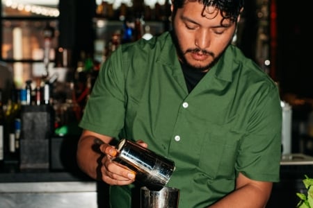 A bartender in a green shirt mixing a craft cocktail behind a warmly lit bar.