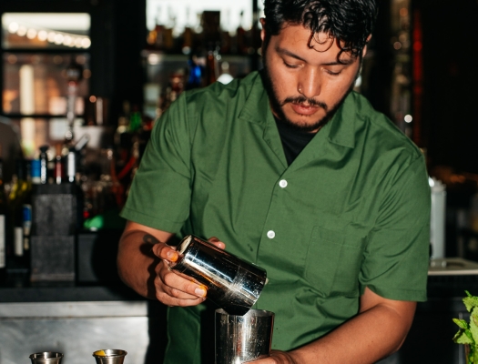 A bartender in a green shirt mixing a craft cocktail behind a warmly lit bar.