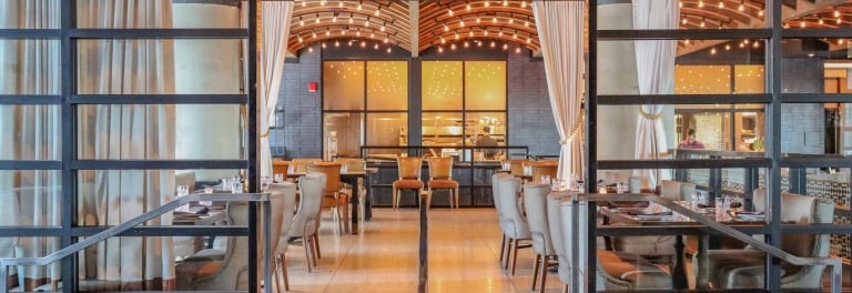 Elegant restaurant interior with vaulted wood ceiling, Edison string lights, and glass-partitioned dining sections with upholstered chairs.
