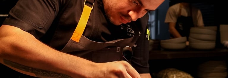 Chef in dark uniform carefully plates a delicate dish on a textured white ceramic bowl in a warmly lit kitchen.