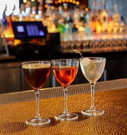 Three cocktails in Nick & Nora glasses on a gold bar top: espresso martini, amber spritz, and clear martini with onions.