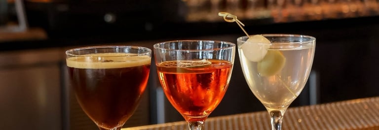 Three cocktails in Nick & Nora glasses on a gold bar top: espresso martini, amber spritz, and clear martini with onions.